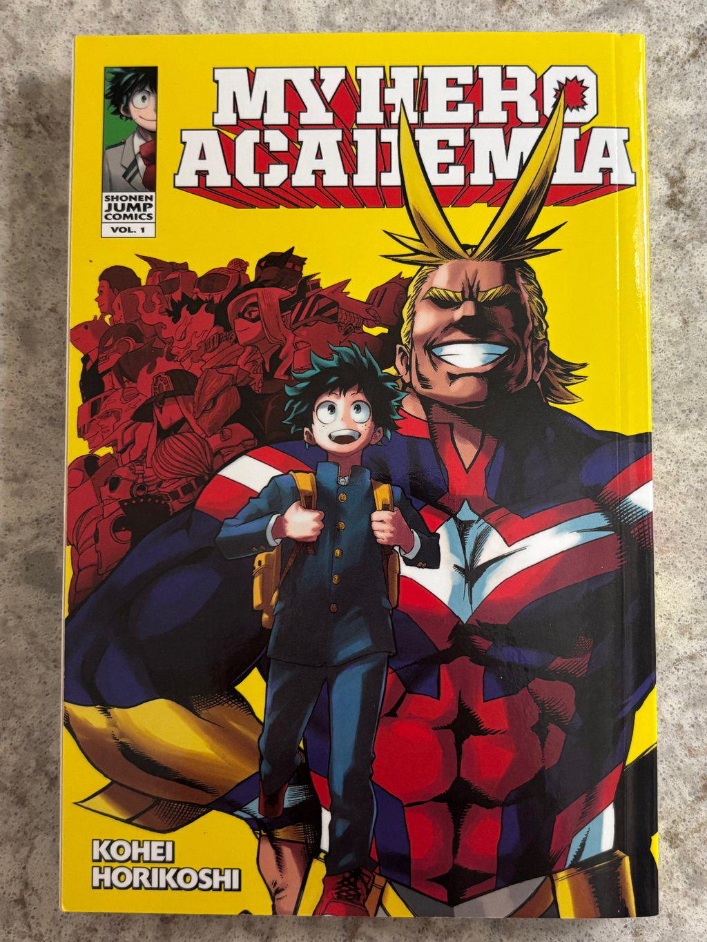 A copy of the My Hero Academia Volume 1 manga book by Kohei Horikoshi, featuring a bright yellow cover with illustrations of Aizawa and Deku.