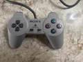 A classic gray Sony PlayStation 1 controller with its attached cable, resting on a textured beige stone surface.